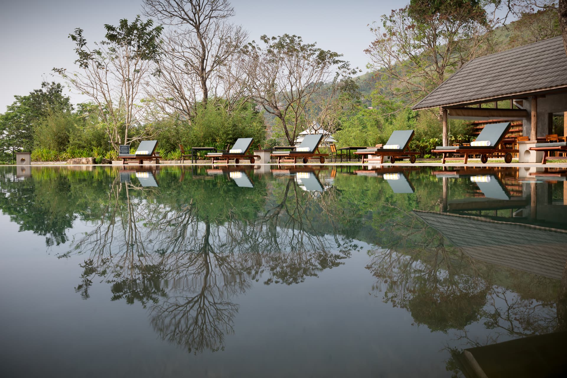 Loungers and pavilion reflected in the calm swimming pool surrounded by trees at Prakriti Shakti
